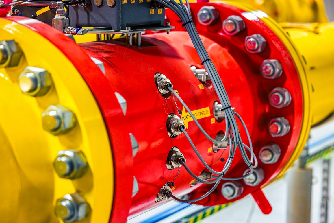 Makro ujęcie nowoczesnego urządzenia pomiarowego wpiętego w rurociąg. Jaskrawożółte kołnierze z wielkimi śrubami spinają czerwony, cylindryczny element z naklejką i strzałką „POSITIVE FLOW DIRECTION”. Do urządzenia podłączone są szare kable.