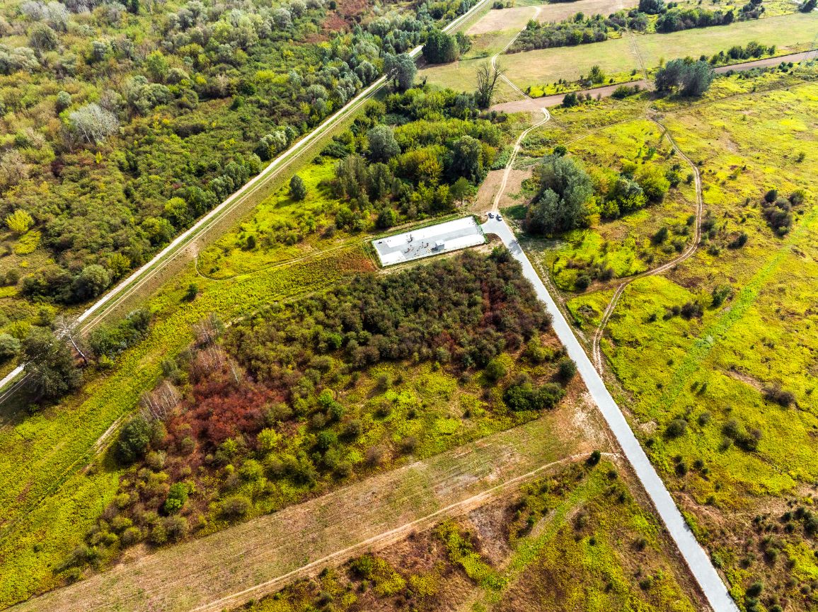 Widok z lotu ptaka na niewielki, ogrodzony, prostokątny plac technologiczny z jasną nawierzchnią tłuczniową, wkomponowany w naturalny krajobraz. Do obiektu z prawego dolnego rogu prowadzi prosta, szara droga dojazdowa. Teren wokół stacji to mozaika zielono-żółtych łąk, pól oraz zagajników z kępami drzew, z których część przybiera już rdzawe, jesienne barwy.