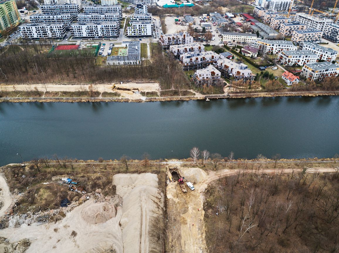 Widok z lotu ptaka na kanał wodny dzielący kadr poziomo. Na górnym brzegu znajduje się nowoczesne osiedle mieszkaniowe z białymi blokami. Na dolnym brzegu widoczny jest plac budowy z usypanymi hałdami piasku, głębokim wykopem ziemnym zabezpieczonym szalunkiem przy samej wodzie oraz zaparkowanym białym busem w sąsiedztwie drzew.