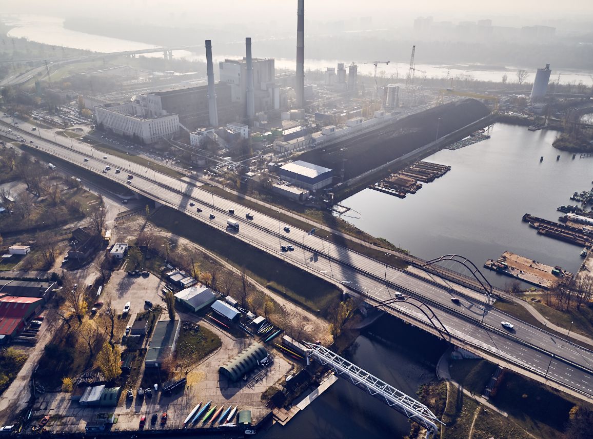 Widok z powietrza w ciągu dnia na duży kompleks przemysłowy elektrociepłowni z wysokimi kominami, spowity lekkim smogiem lub mgłą. Na pierwszym planie szeroki, wielopasmowy most drogowy z jadącymi samochodami przecina rzekę. Poniżej mostu widoczna jest mniejsza, biała kładka łukowa oraz baza zadaszonych kajaków przy brzegu.