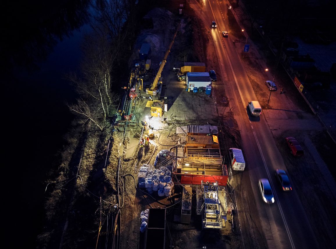 Nocne zdjęcie z lotu ptaka przedstawiające plac budowy zlokalizowany tuż obok oświetlonej drogi, po której poruszają się samochody. Teren prac jest jaskrawo rozświetlony reflektorami; widać na nim ciężki sprzęt budowlany (w tym palownicę i dźwig), kontenery pracownicze oraz ułożone materiały konstrukcyjne nad brzegiem ciemnej wody.