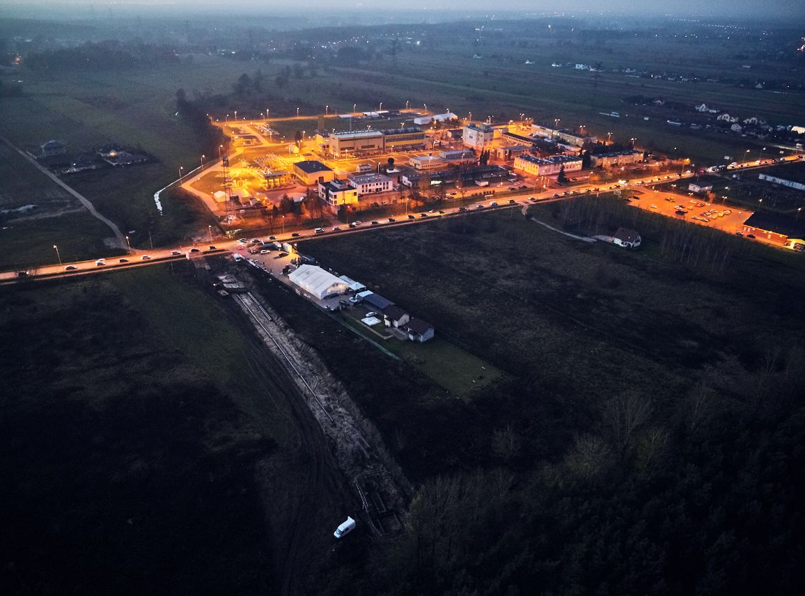 Szerokie ujęcie z powietrza wykonane o zmierzchu. W centralnej części widoczna jest nowoczesna, silnie oświetlona żółtym światłem tłocznia gazu lub stacja przesyłowa, otoczona ciemnymi polami. W dolnej części kadru, od strony lasu, ciągnie się podłużny wykop budowlany z pracującym sprzętem i białym pojazdem technicznym.