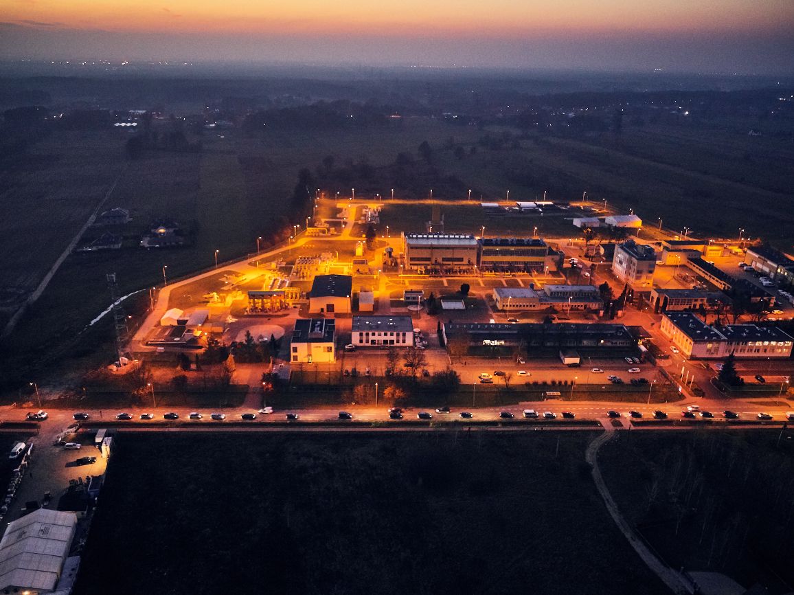 Zdjęcie lotnicze z bliskiej odległości przedstawiające oświetloną nocą instalację przemysłową gazociągu. Kompleks budynków, rurociągów i urządzeń technicznych jarzy się jasnym, żółto-pomarańczowym światłem. Na dole kadru przebiega prosta droga, na której reflektory samochodów tworzą jasny sznur świateł.