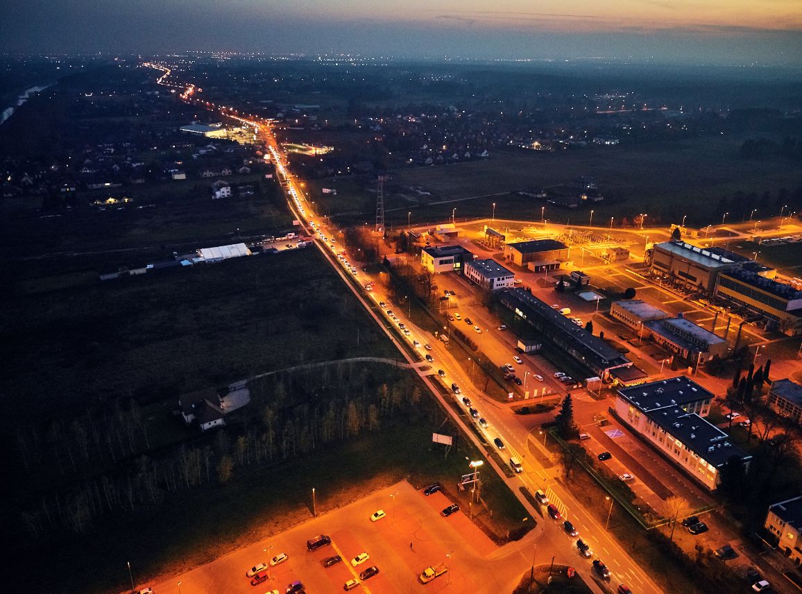 Nocna panorama z powietrza ukazująca infrastrukturę przemysłową w otoczeniu podmiejskim. Przez kadr ukośnie przebiega długa ulica rozświetlona latarniami i reflektorami aut. Po prawej stronie znajduje się jasny, iluminowany zakład techniczny, natomiast na pierwszym planie widoczny jest duży parking oświetlony ciepłym, pomarańczowym blaskiem.