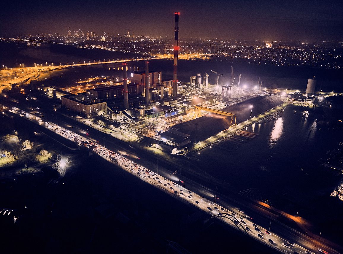 Nocny widok z lotu ptaka na potężny kompleks elektrociepłowni Żerań. Obiekt jest gęsto oświetlony, a jego wysokie kominy posiadają czerwone światła ostrzegawcze. Na pierwszym planie widać estakadę i most z intensywnym ruchem samochodowym, którego światła tworzą ciągłe linie. W tle majaczą rozproszone światła nocnej Warszawy.