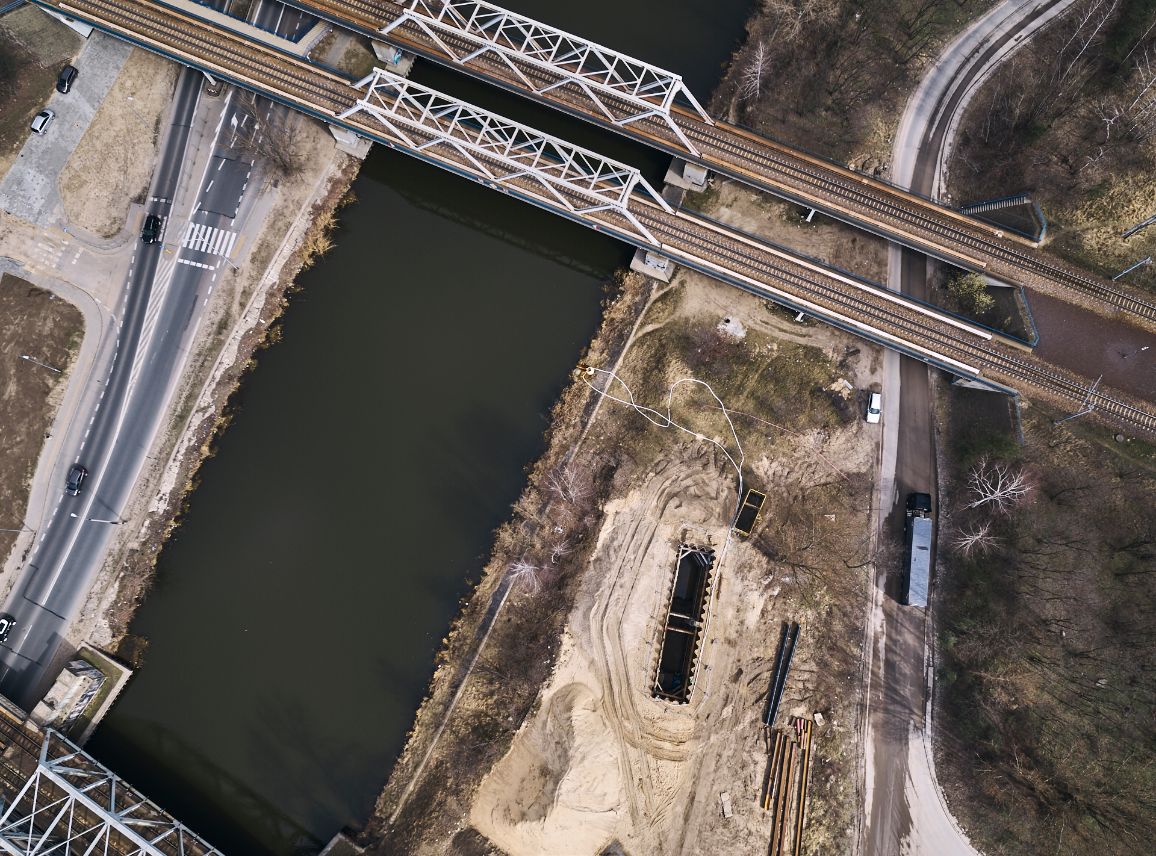 Widok z góry (pionowo) na infrastrukturę transportową nad kanałem wodnym. Górną część kadru przecinają dwa równoległe, stalowe mosty kolejowe o kratownicowej konstrukcji. Na prawym brzegu, w pasie zieleni między torami a drogą, znajduje się głęboki, prostokątny wykop budowlany zabezpieczony ściankami szczelnymi (larssenami), do którego doprowadzono białe rury instalacyjne.