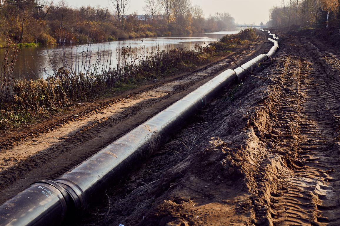 Długi ciąg zespawanych, czarnych rur gazowych ułożony na piaszczystym nasypie wzdłuż koryta rzeki. Rura ciągnie się łukiem wzdłuż brzegu aż po horyzont. Na ziemi widoczne są głębokie ślady opon maszyn budowlanych. Po drugiej stronie rzeki rosną bezlistne, jesienne drzewa.
