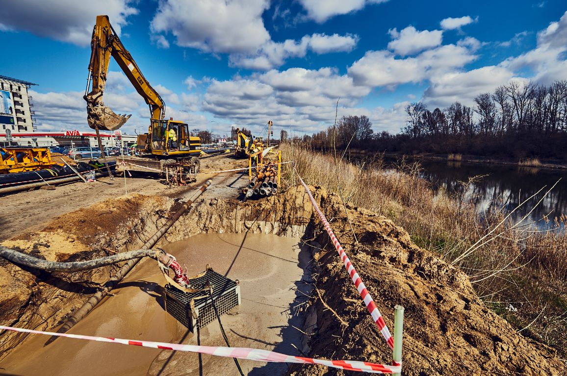 Plac budowy nad brzegiem rzeki w słoneczny dzień z dynamicznymi chmurami. Na pierwszym planie znajduje się głęboki dół wypełniony błotnistą wodą, z którego pompa odprowadza ciecz za pomocą grubego węża. Teren zabezpieczono biało-czerwoną taśmą ostrzegawczą. W tle pracuje żółta koparka gąsienicowa marki CAT, a w oddali widać stację paliw Avia.