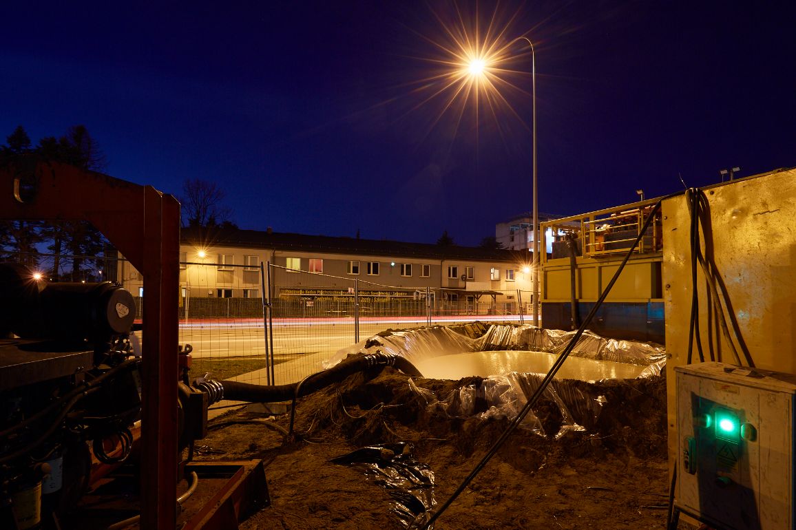 Plac budowy gazociągu nocą, oświetlony mocnym światłem latarni ulicznej dającym efekt gwiazdy. Na pierwszym planie widoczny jest fragment czerwonej konstrukcji maszyny oraz wykop zabezpieczony grubą, przezroczystą folią, w którym znajduje się jasna substancja lub woda. W tle, za metalowym płotem, stoi oświetlony budynek mieszkalny z widoczną tablicą informacyjną.