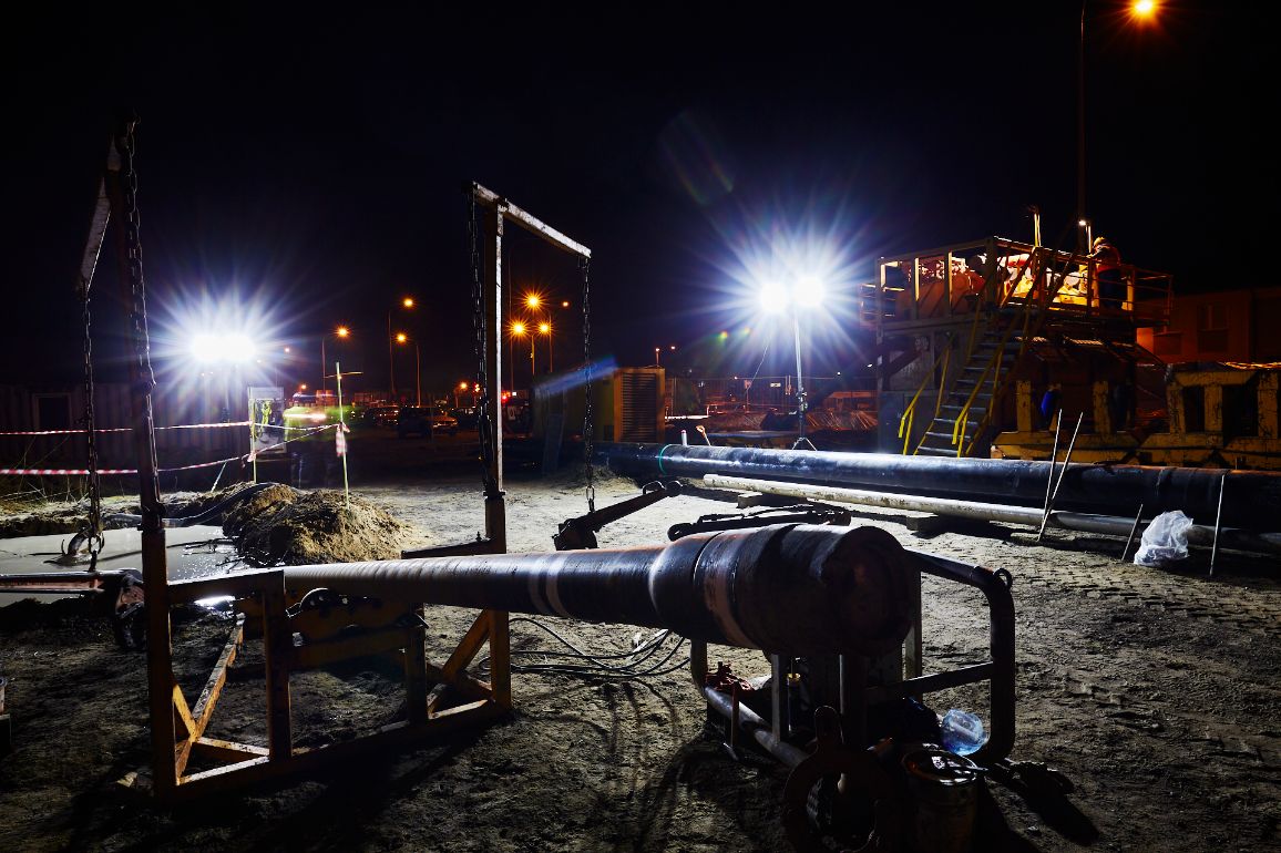 Prace budowlane prowadzone nocą w intensywnym świetle reflektorów budowlanych. Na piaszczystym podłożu zamontowano ciężki, metalowy sprzęt do przewiertów sterowanych (HDD) z grubą, metalową żerdzią celującą w grunt. W tle widać ułożone rurociągi, platformy techniczne oraz rozmyte światła przejeżdżających w oddali samochodów.