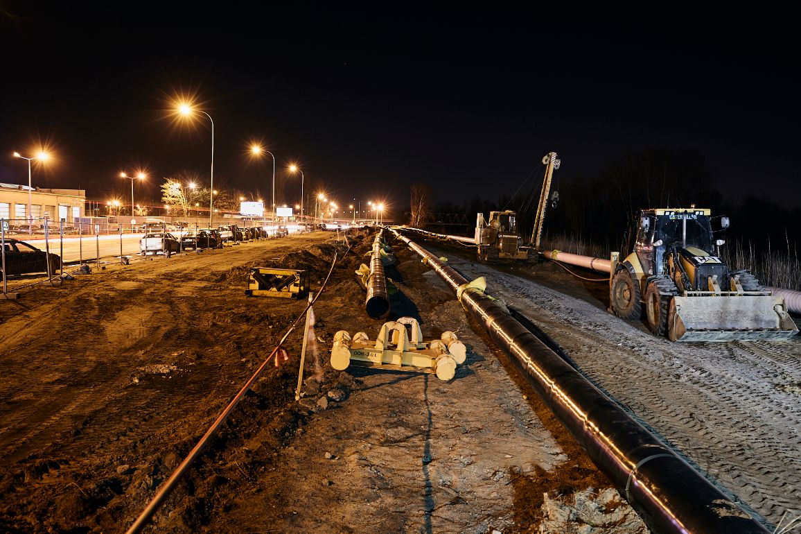 Nocny widok na plac budowy wzdłuż drogi oświetlonej żółtymi latarniami ulicznymi. Przez środek kadru biegnie długi, błyszczący ciąg czarnych rur gazowych ułożonych na piaszczystym pasie. Po prawej stronie stoi żółto-czarna koparko-ładowarka marki CAT, a na ziemi leżą żółte pasy transportowe i elementy montażowe.